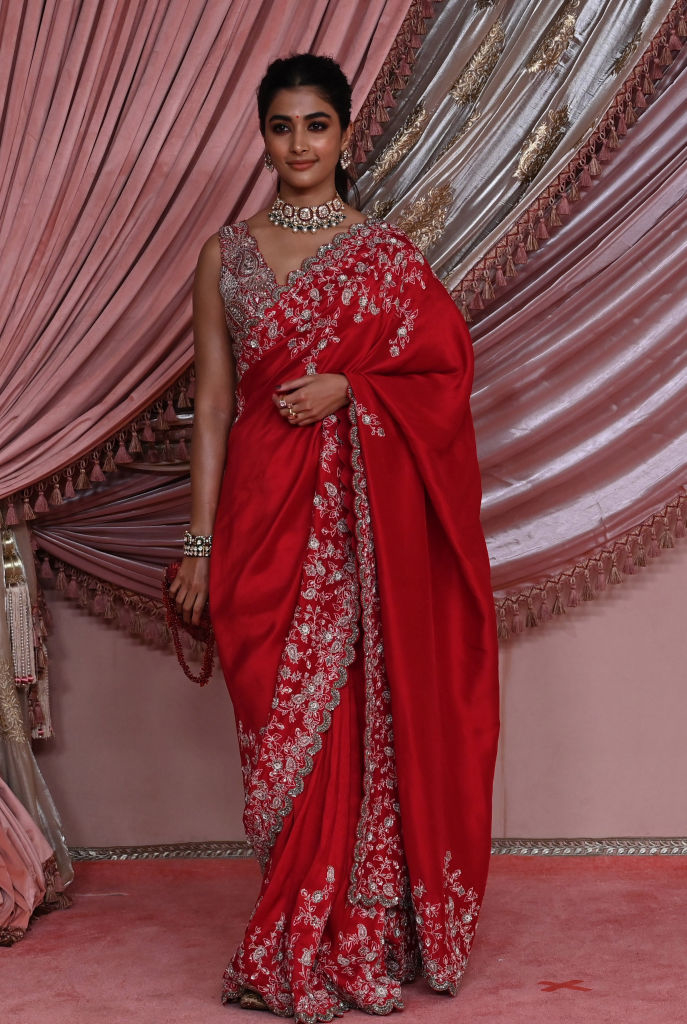 Pooja Hegde stands in front of a draped backdrop, wearing an ornate sari with intricate detailing and a statement necklace