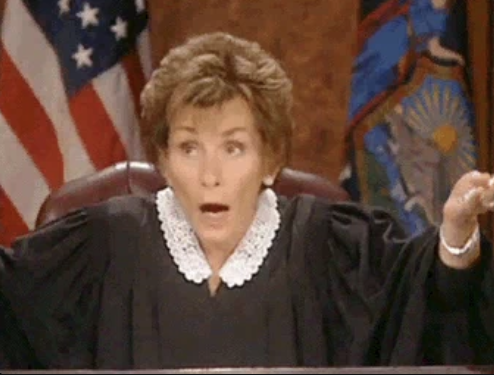Judge Judy in her courtroom, wearing a traditional judge’s robe with a white lace collar, raises her arms in surprise or disbelief