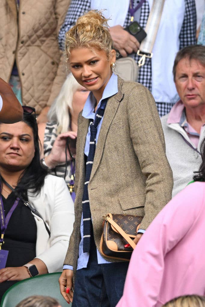 Zendaya stands in a crowd wearing a light shirt, striped tie, and a textured blazer, with a crossbody bag and red nail polish