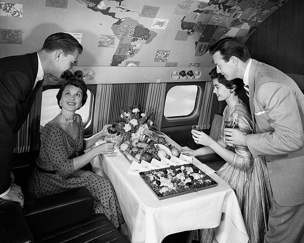 Nancy Kelly, seated, shares a meal with Michael Wilding and Elizabeth Taylor; a male attendant stands nearby in an elegantly decorated airplane cabin
