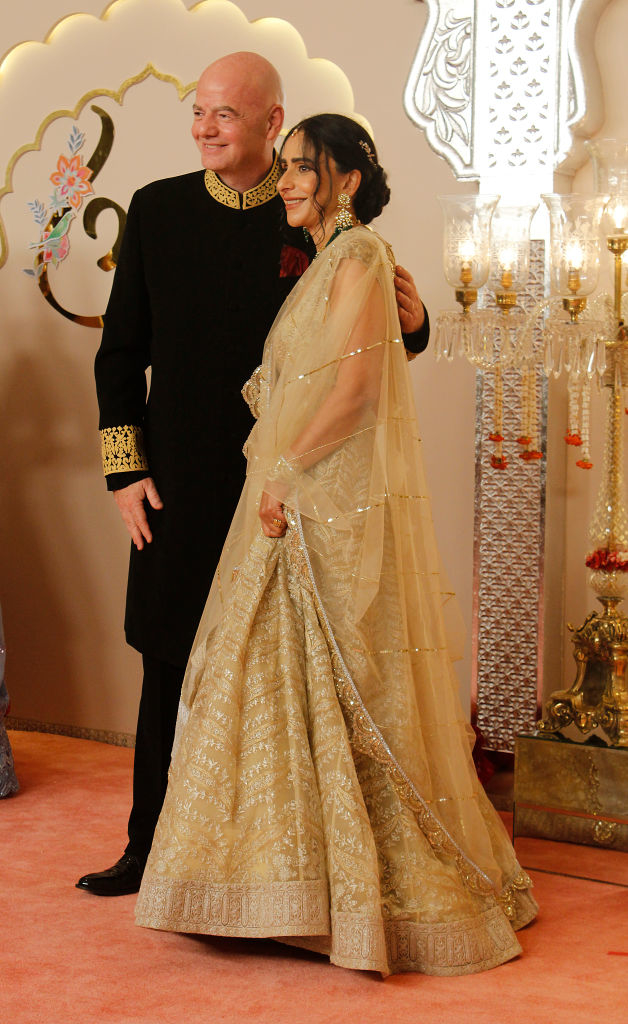 Gianni in an embroidered outfit poses with Leena in a detailed, ornate gown and veil at a formal event