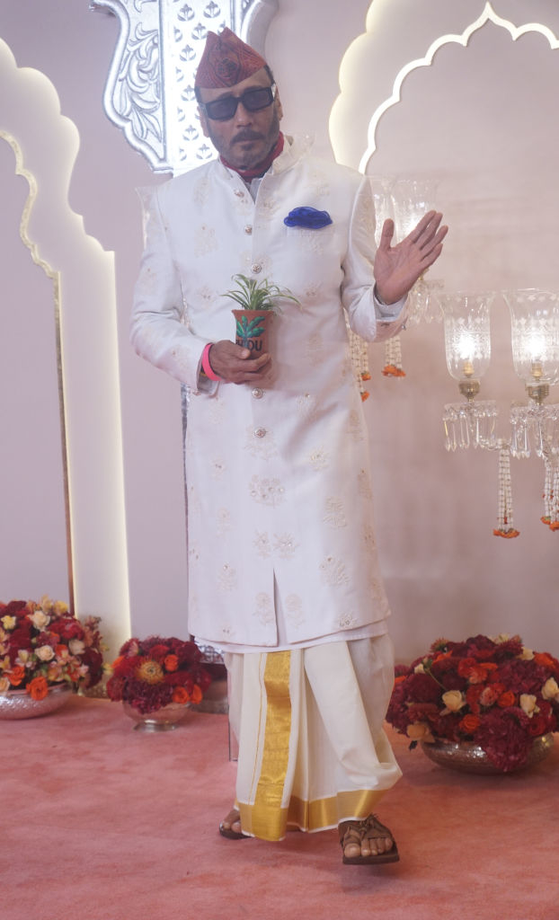 Jackie wearing a sherwani and dhoti with shiny trim. He is holding a small plant and standing on a stage with floral decorations and chandeliers