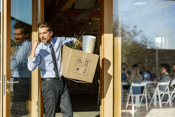A man in business attire, carrying a box with office items, happily exits a building while people sit in a meeting room in the background