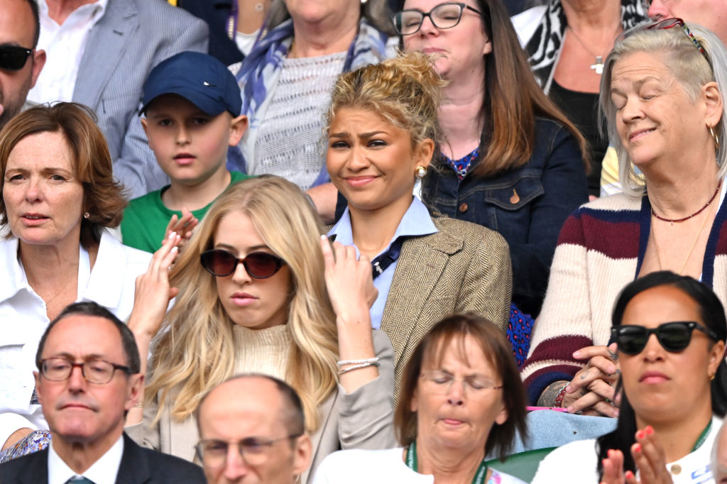 Zendaya sitting in a crowded audience, wearing a blue shirt with a blazer. Other people in the image include a blonde woman in sunglasses and various adults looking on