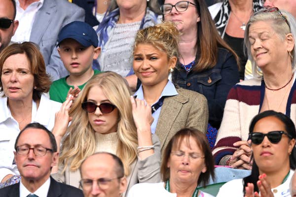 Zendaya sitting in a crowded audience, wearing a blue shirt with a blazer. Other people in the image include a blonde woman in sunglasses and various adults looking on