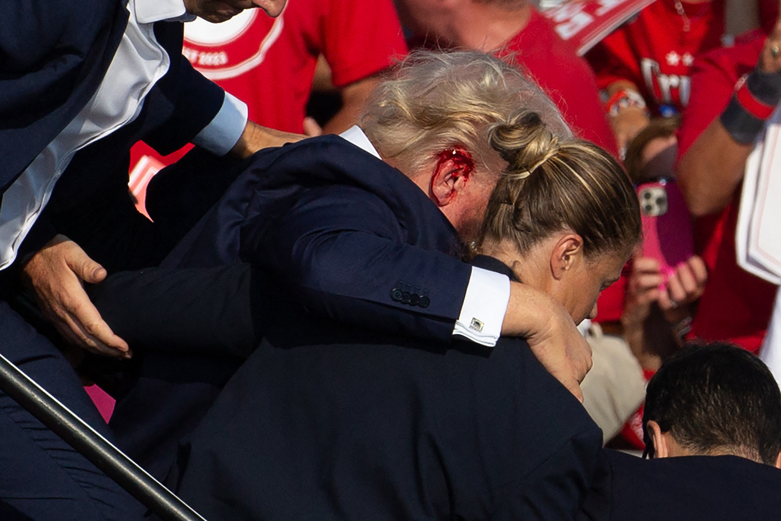 Donald Trump, with a bleeding head wound, is supported by a woman and others at an event