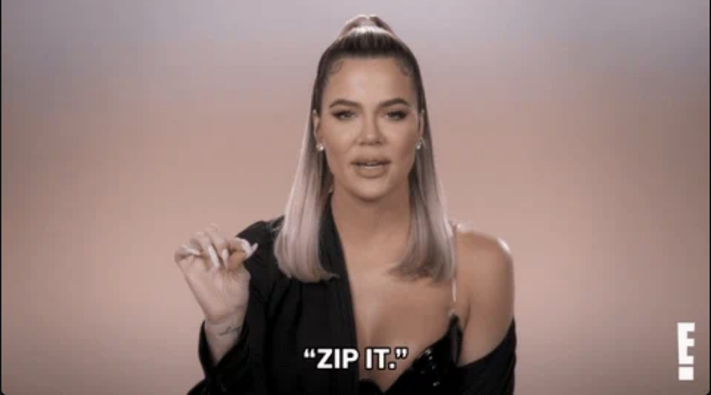 Khloe Kardashian wearing a stylish, off-shoulder outfit, gestures to &quot;zip it&quot; with her hand. Text reads &quot;ZIP IT.&quot; in the image