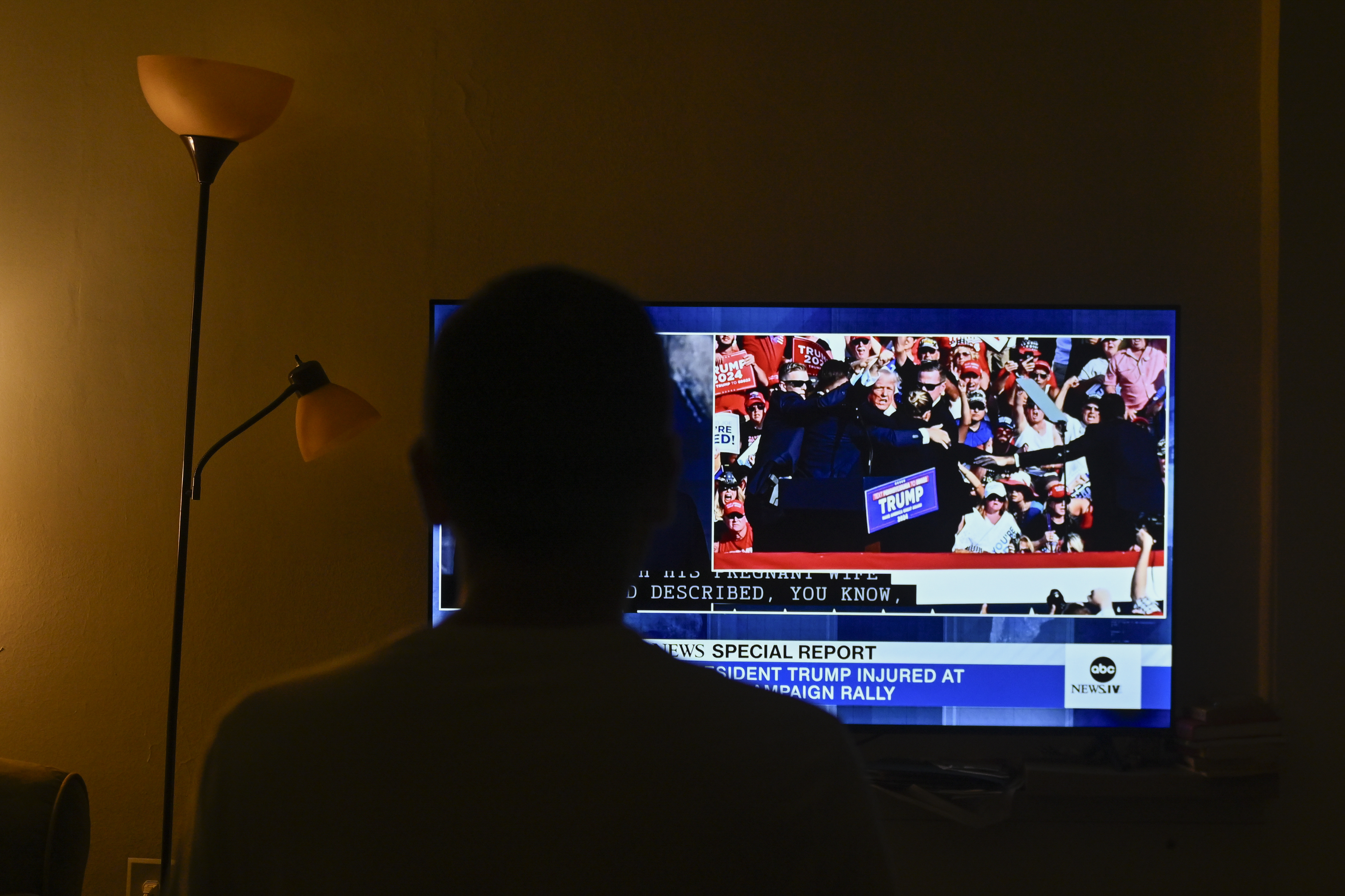 A person watches a news report on TV about an incident at a President Trump campaign rally, showing a crowd and Trump on stage