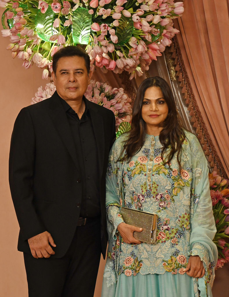 Atul in dark suit and Alvira in embroidered dress stand together in front of a flower arrangement