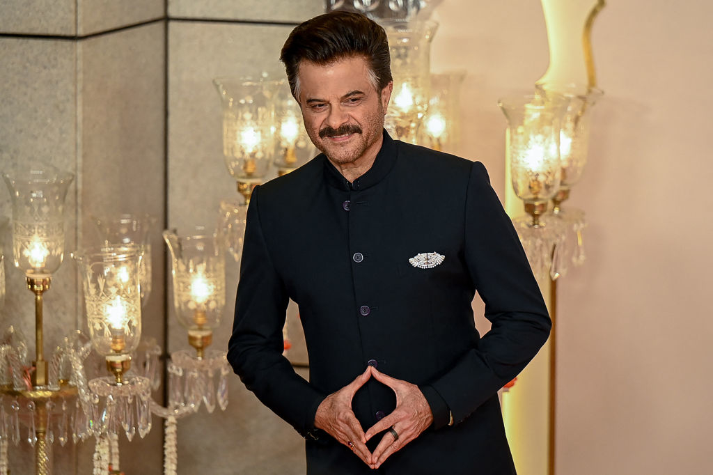 Anil Kapoor poses on the red carpet in an elegant, dark tailored suit with a decorative pocket square. He stands in front of ornate light fixtures