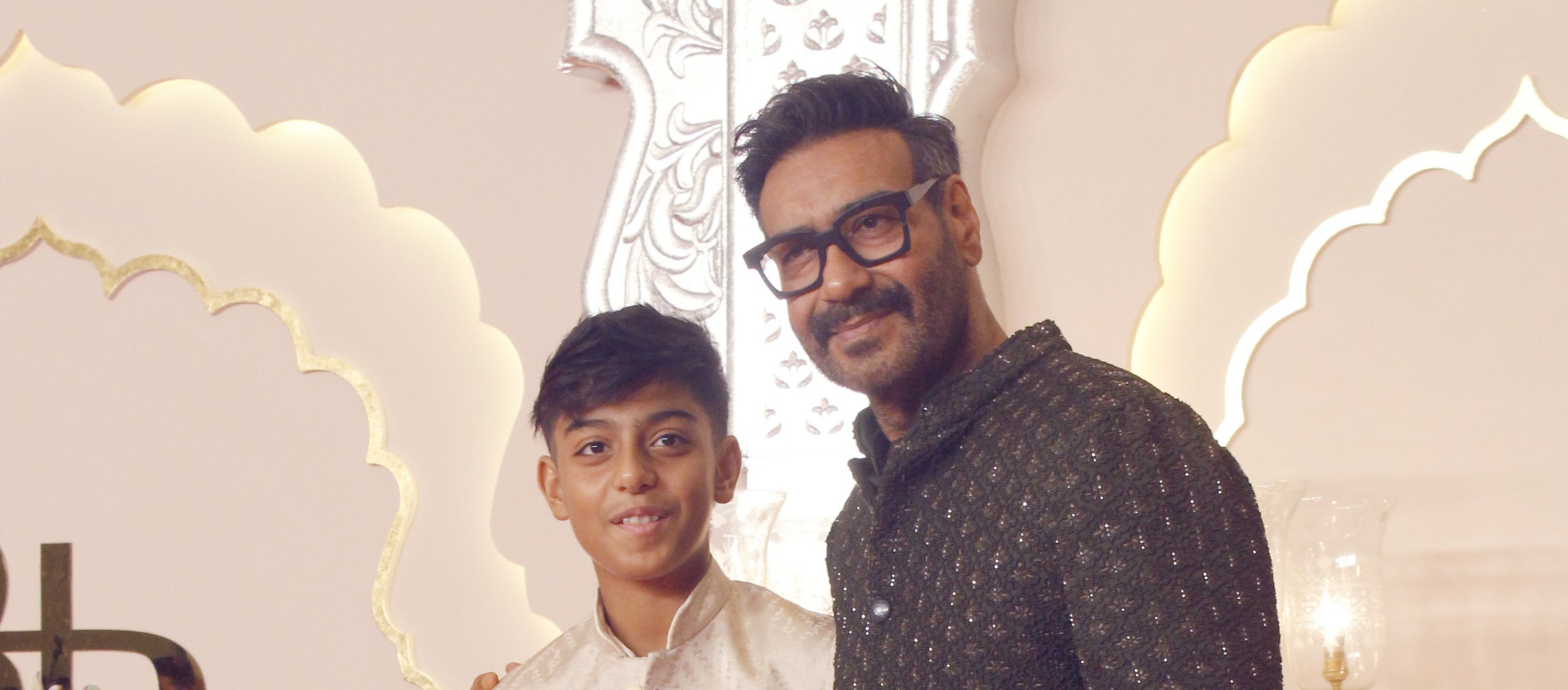 Ajay Devgn with his son Yug Devgn at an event, both wearing traditional Indian attire. Ajay in a dark sherwani, and Yug in a light-colored sherwani