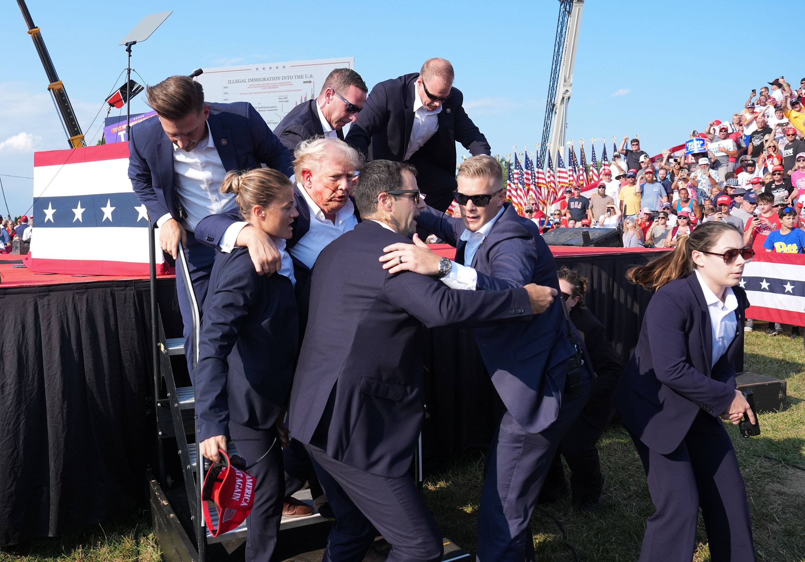 Donald Trump, surrounded by Secret Service agents in suits, being helped off stage at an outdoor rally with supporters in the background