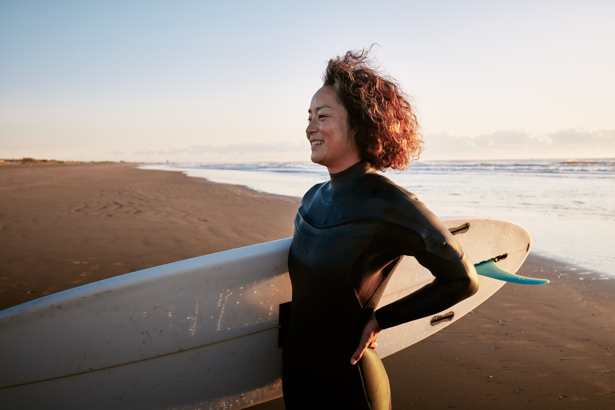 A woman wearing a wetsuit stands on a beach holding a surfboard, looking away from the ocean with a contemplative expression