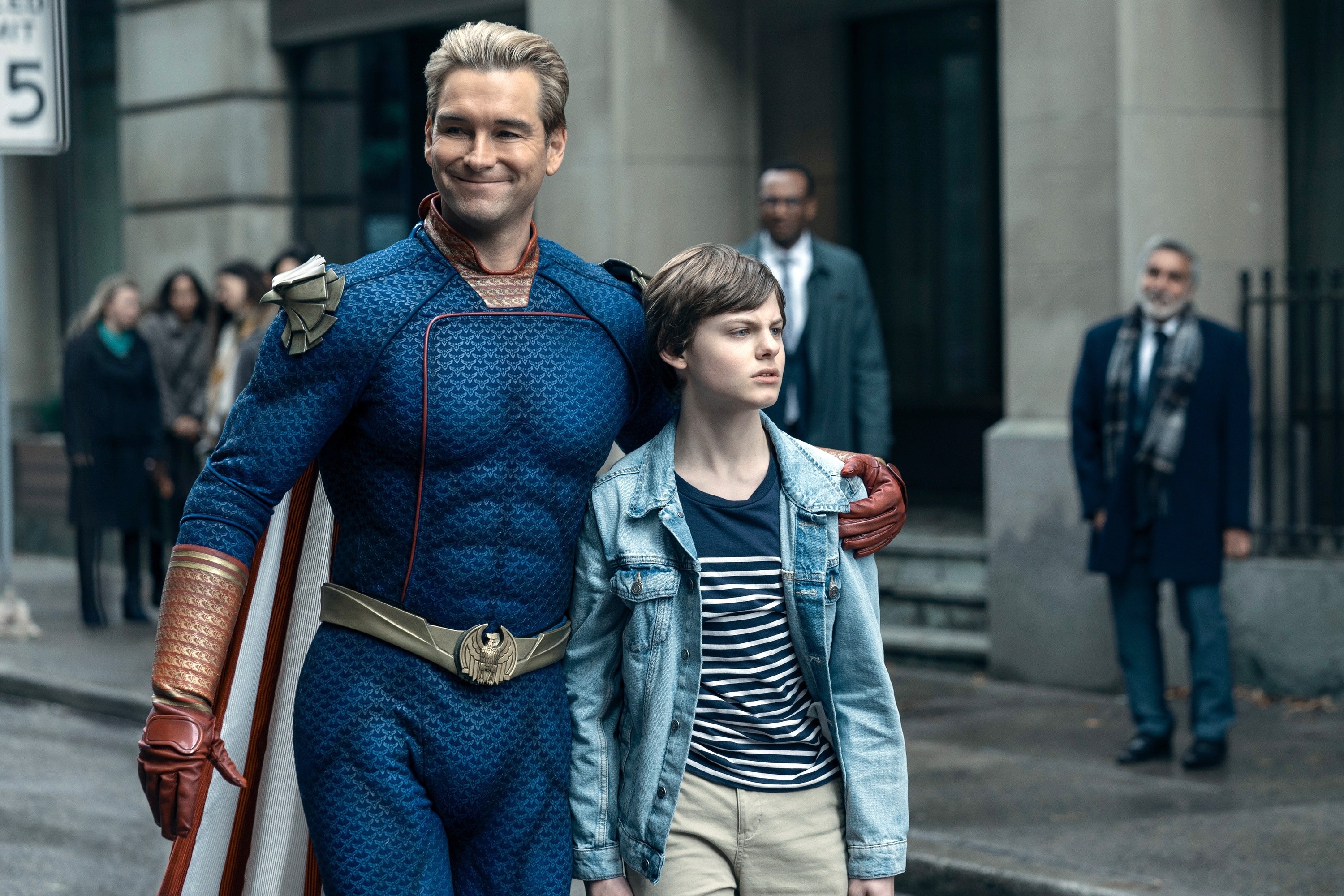 Antony Starr as Homelander in costume, smiling with his arm around Cameron Crovetti as Ryan Butcher, walking on a city street