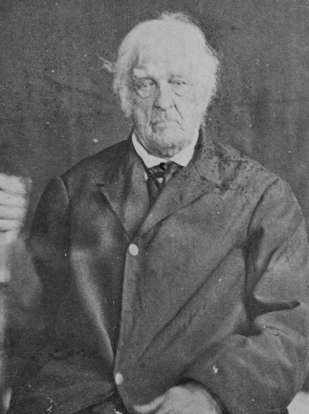An elderly man with thinning hair and a solemn expression wearing a dark coat