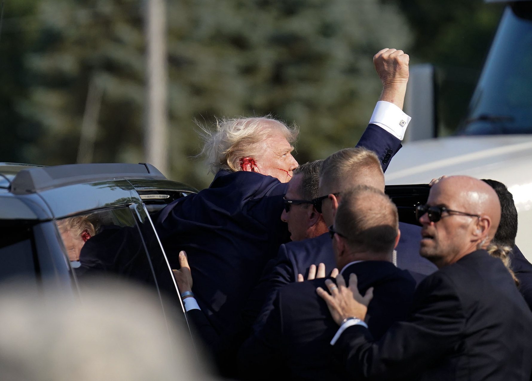 Donald Trump surrounded by security personnel, raising a fist in the air as he enters a vehicle