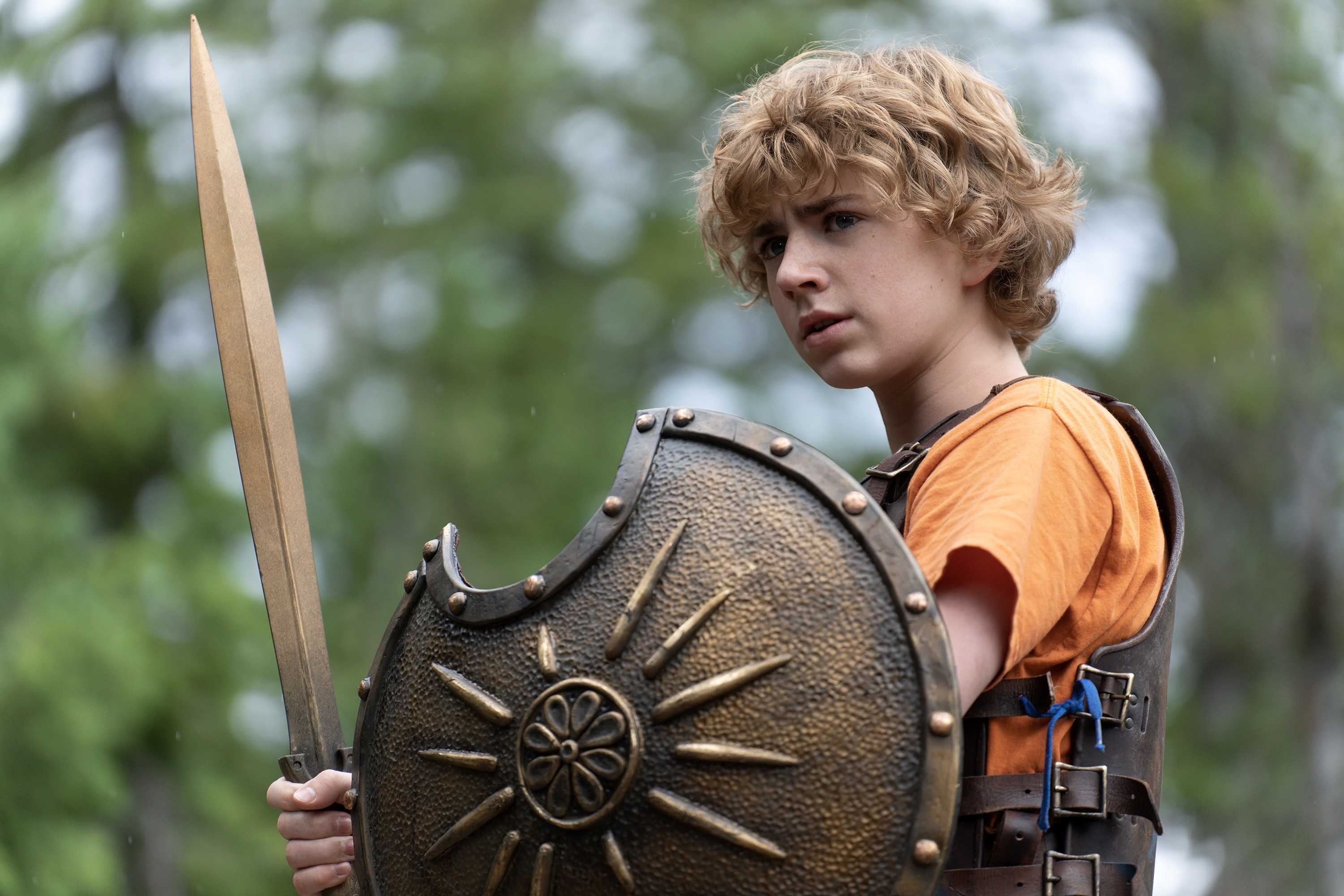 Walker Scobell, dressed as Percy Jackson, holds a sword and shield in an outdoor scene from a TV show