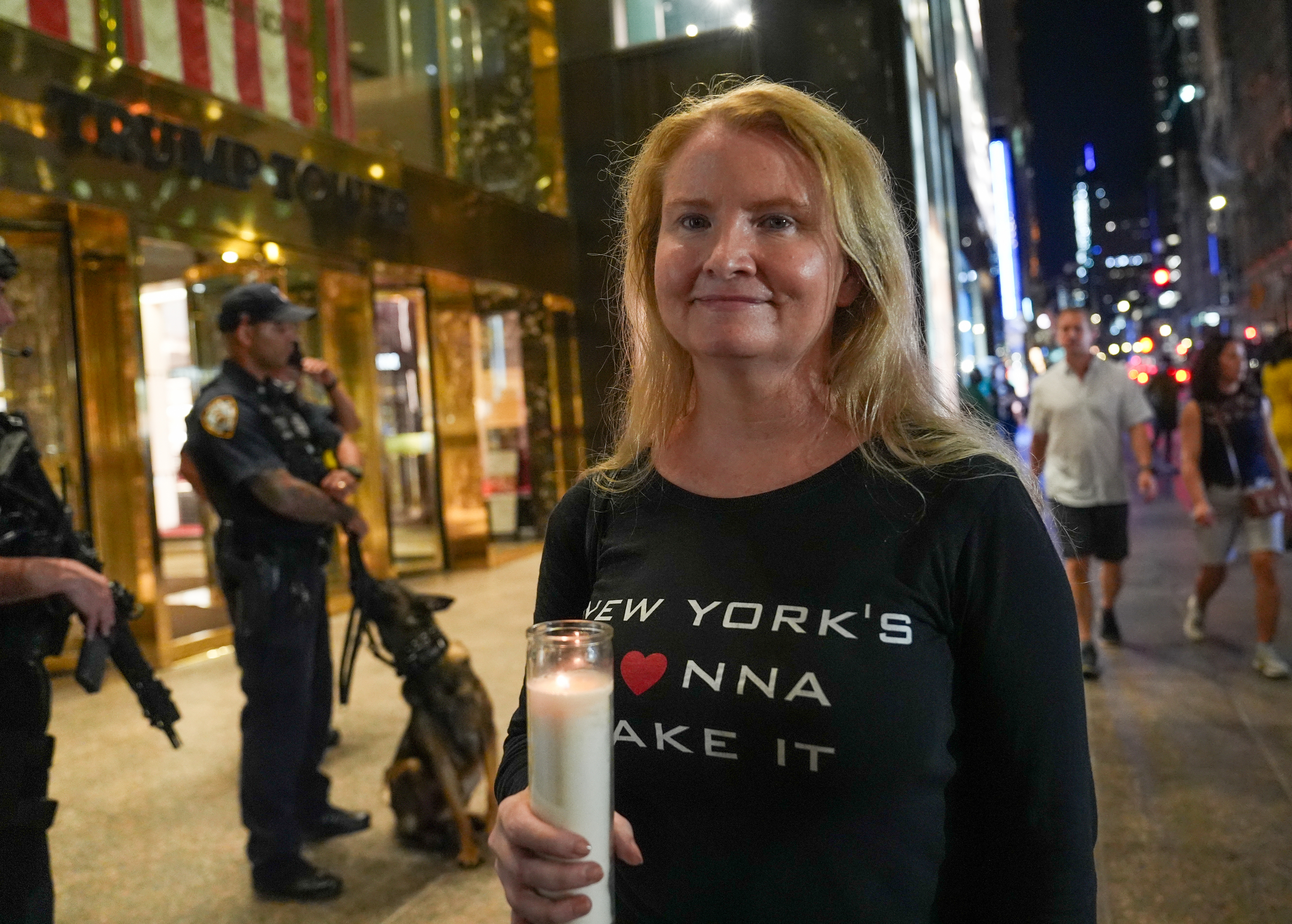 A woman holds a candle on a city street at night. Police officers and a dog stand in the background near a building with a large flag inside