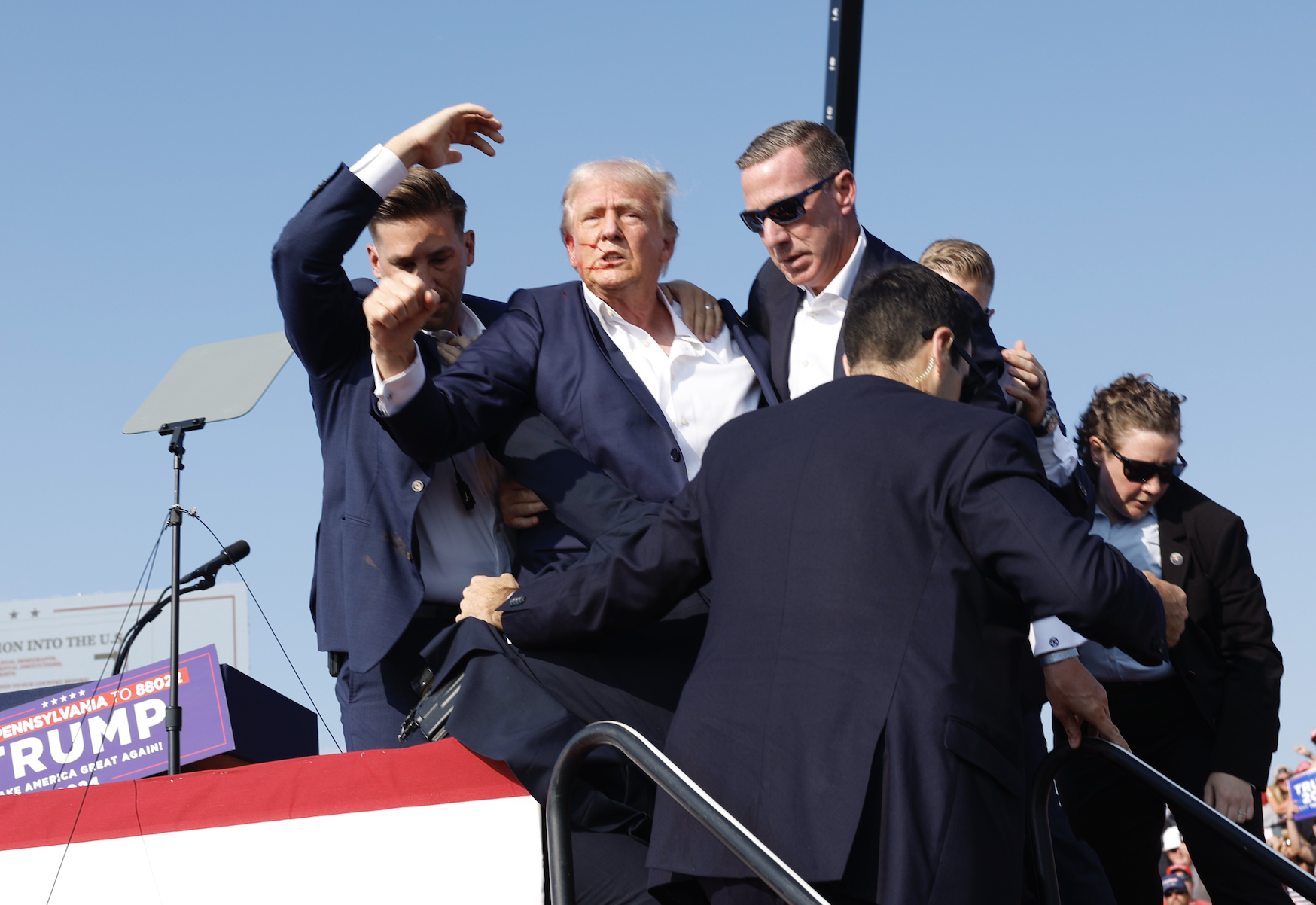 Donald Trump being assisted on stage by several men during a public event. A podium with a sign for his rally is visible