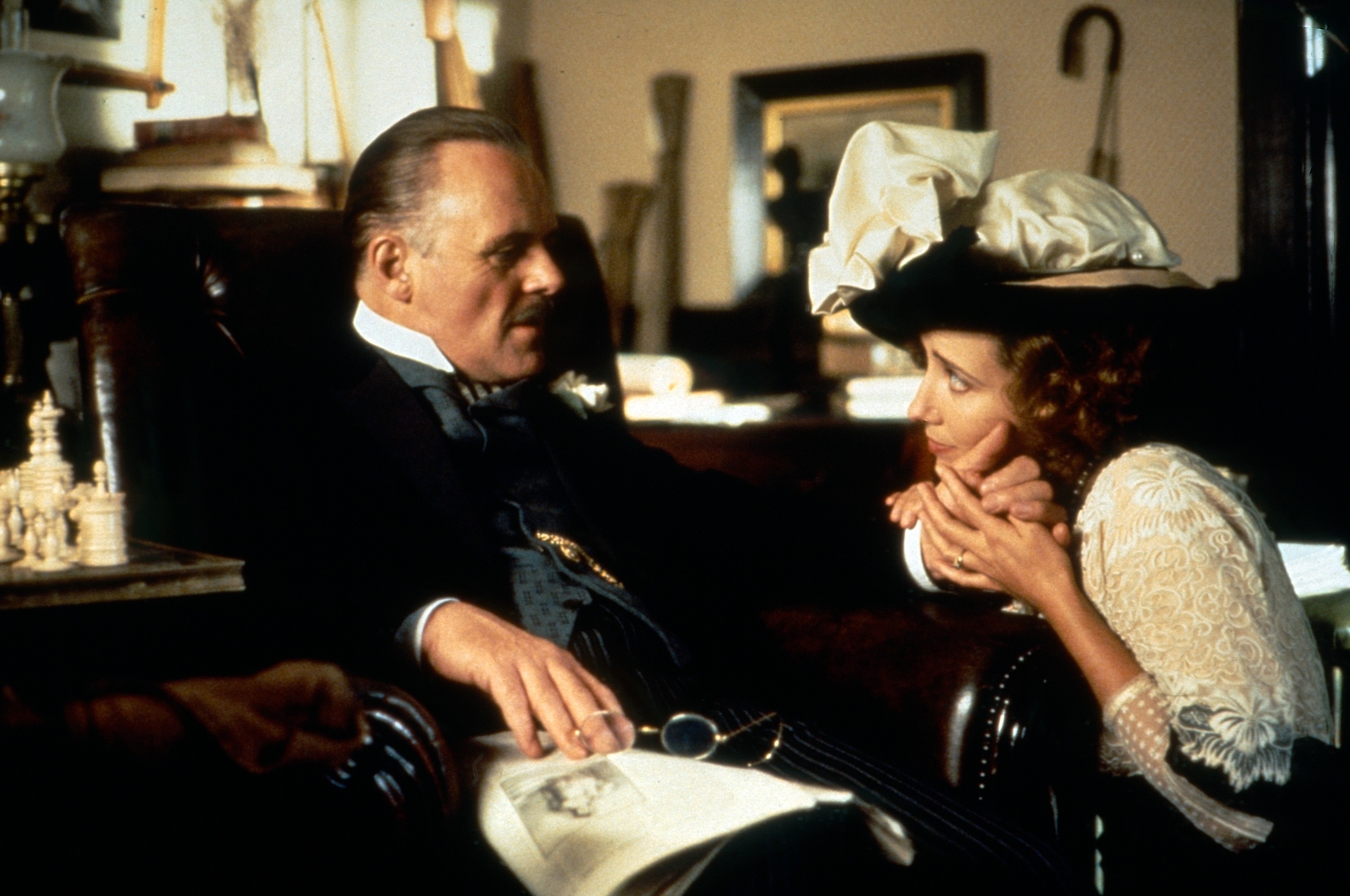 Anthony Hopkins, dressed in a traditional suit, sits in a chair, while Emma Thompson, dressed in period attire with a large hat, holds his hand and rests her cheek on it