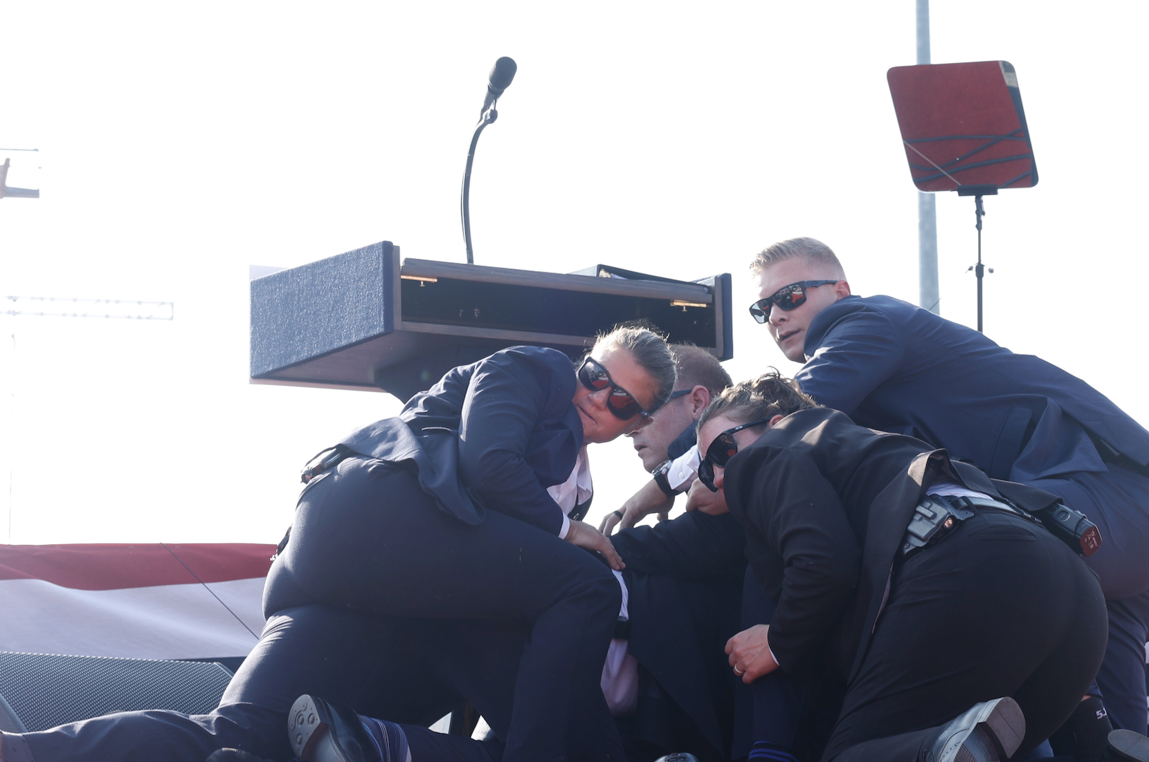 Four security agents in suits and sunglasses use their bodies to shield someone on the ground near a podium with a microphone