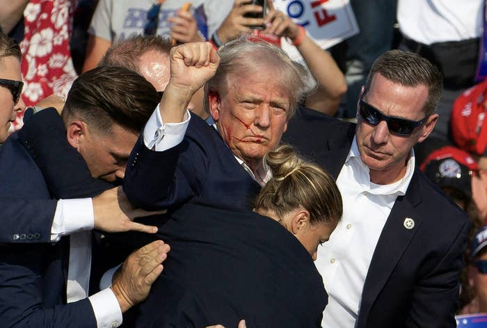 Donald Trump, surrounded by security personnel, raises his fist at an outdoor event. Markings are visible on his face. Other people in suits are assisting