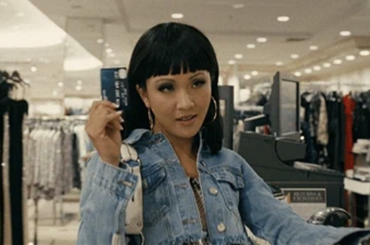 A woman in a denim jacket holds a credit card while shopping in a clothing store. Multiple racks of clothes are visible in the background