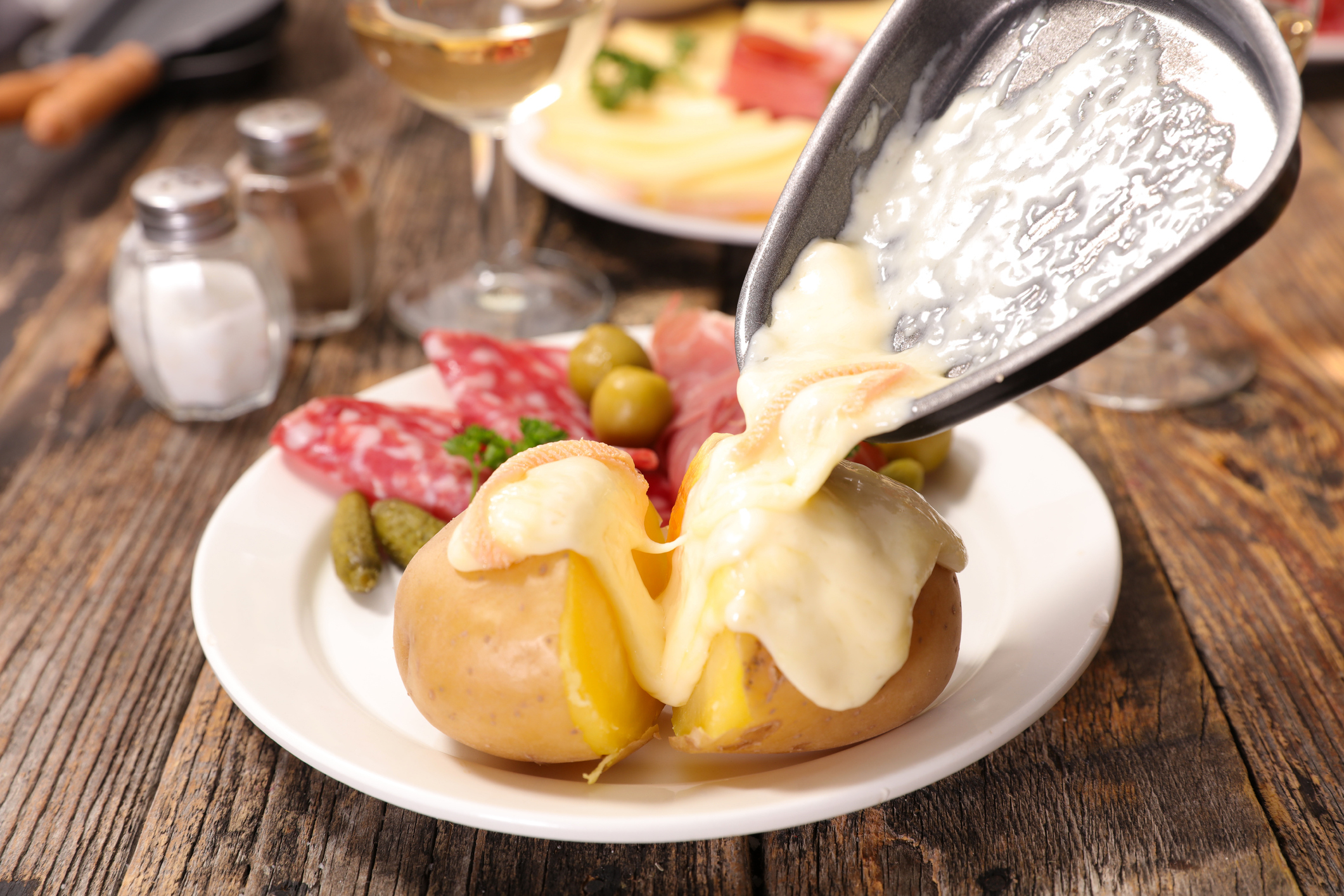 Melted cheese being poured over a baked potato on a plate with olives, pickles, and sliced cured meats