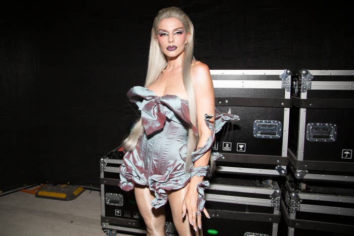Julia Fox stands in front of equipment cases, wearing a dramatic, sculptural dress with large ruffled details. Their long hair is styled straight, and they have dark lipstick