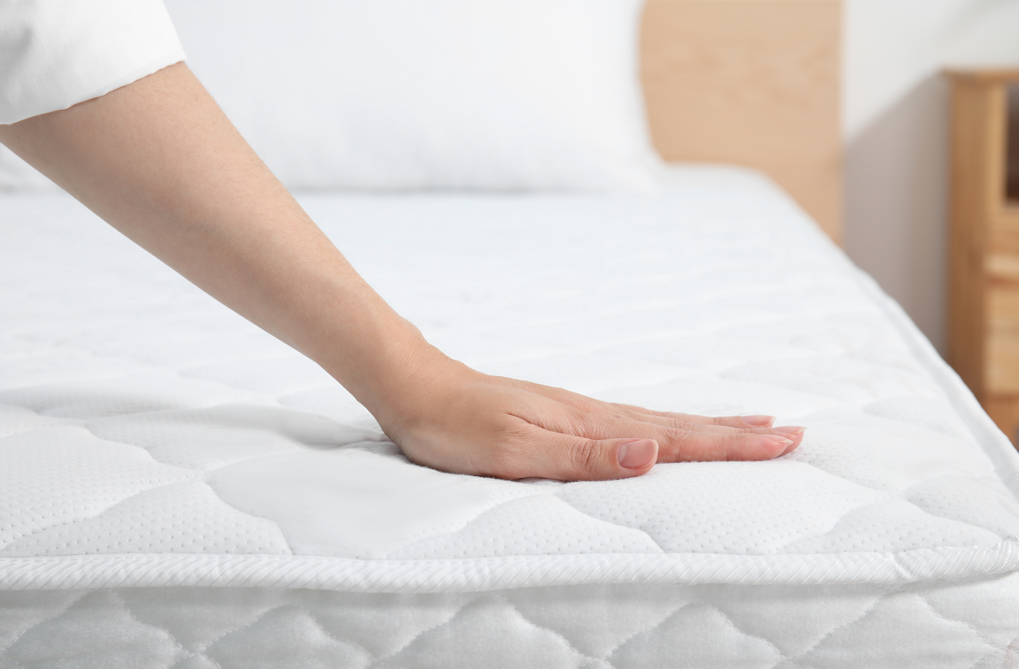 A hand presses down on a mattress in a bedroom, testing its firmness and quality