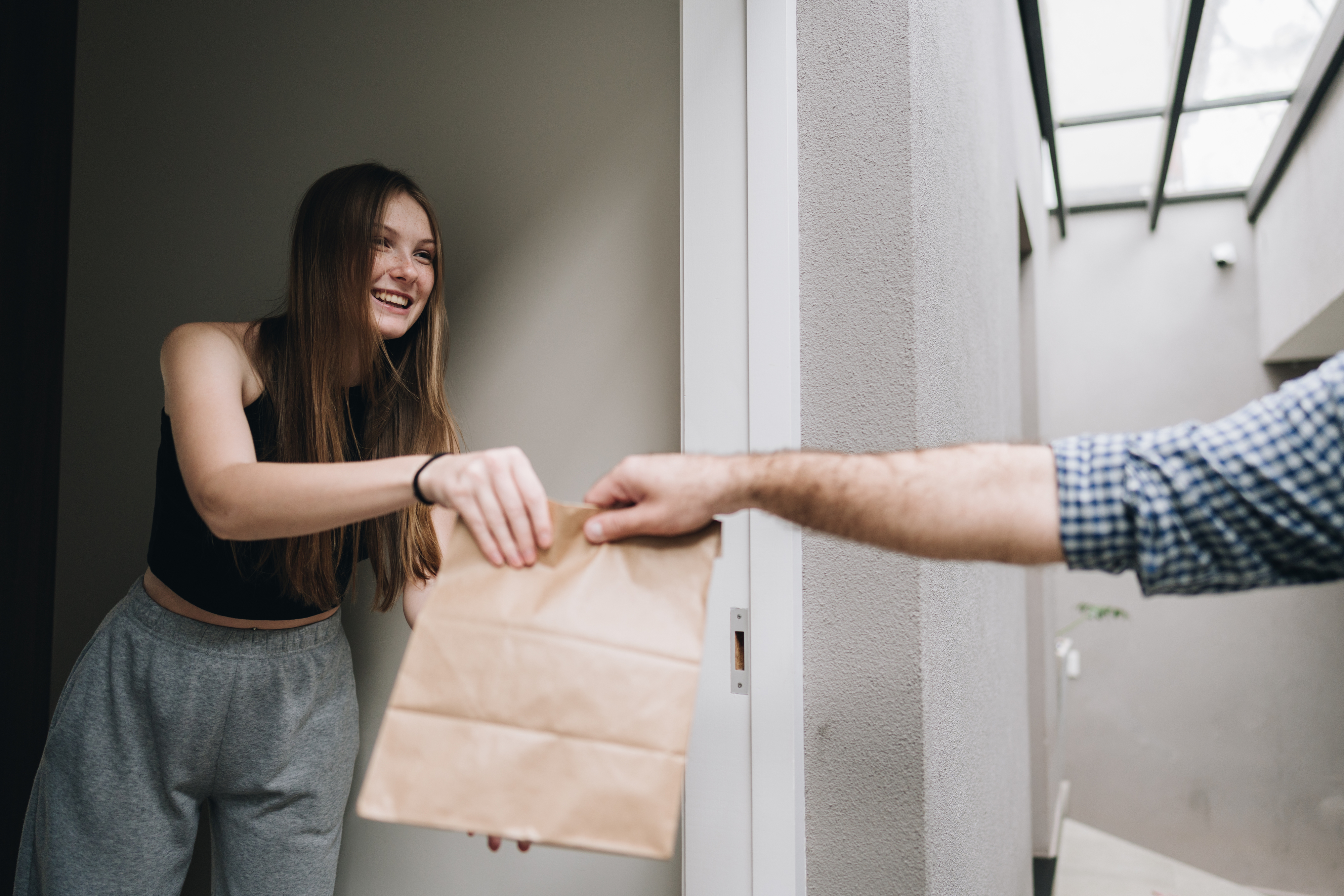 Woman in casual attire happily receives a brown paper bag from a person at the doorway of a modern building