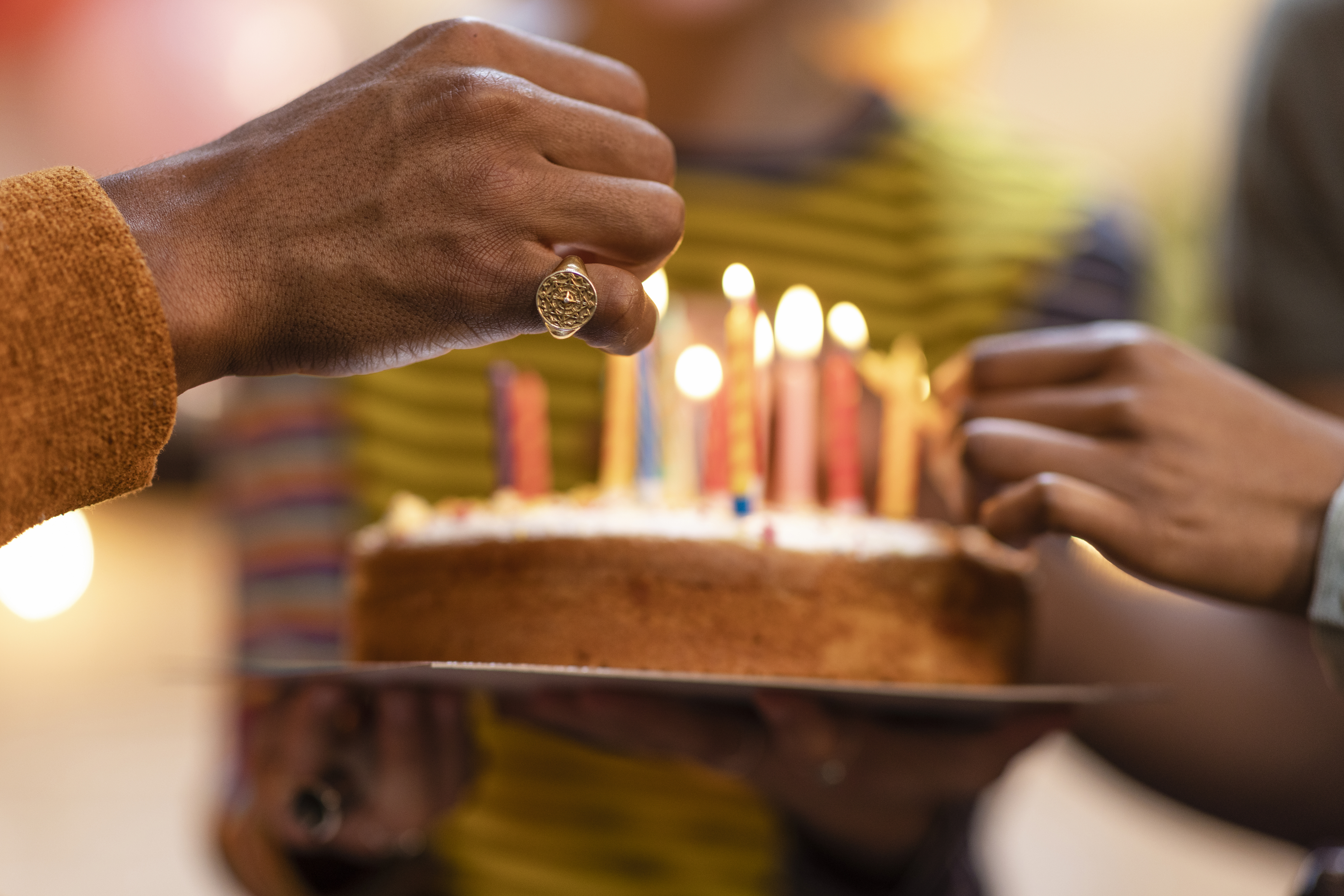 A close-up of a birthday cake with lit candles, with two unidentified people holding the cake and lighting the candles
