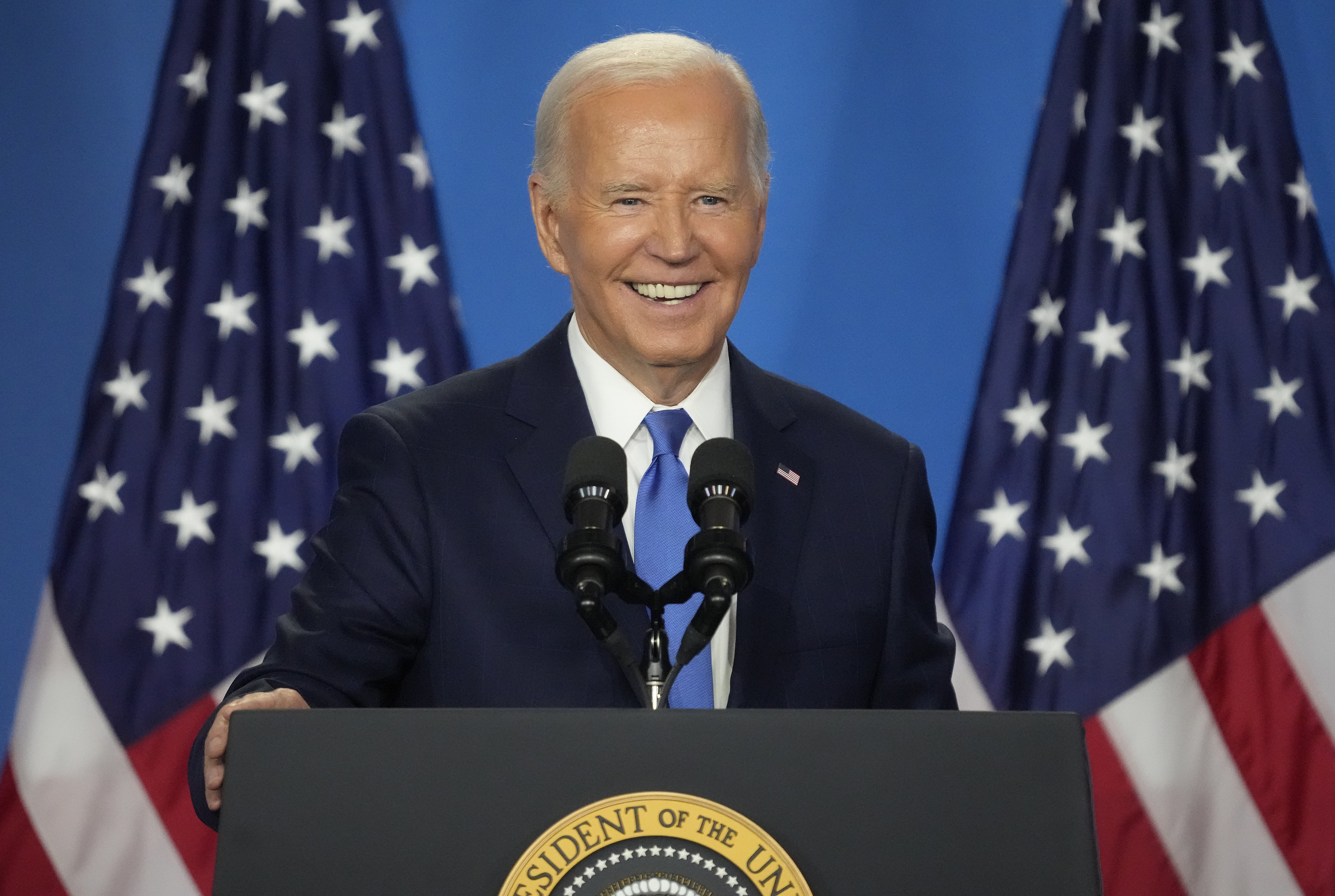 Joe Biden speaks at a podium, smiling, with American flags in the background