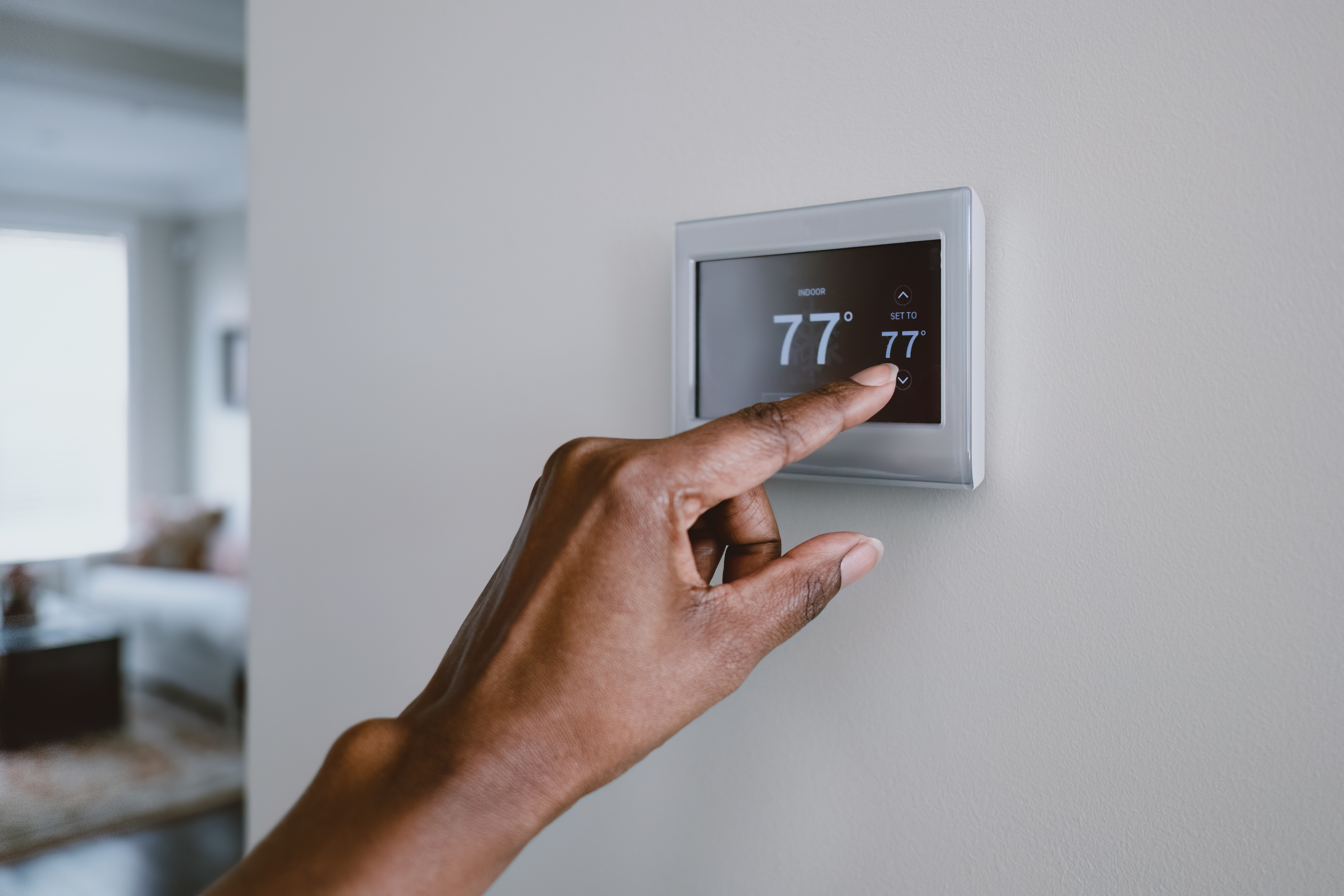 A hand adjusts a digital thermostat on the wall, setting it to 77 degrees Fahrenheit