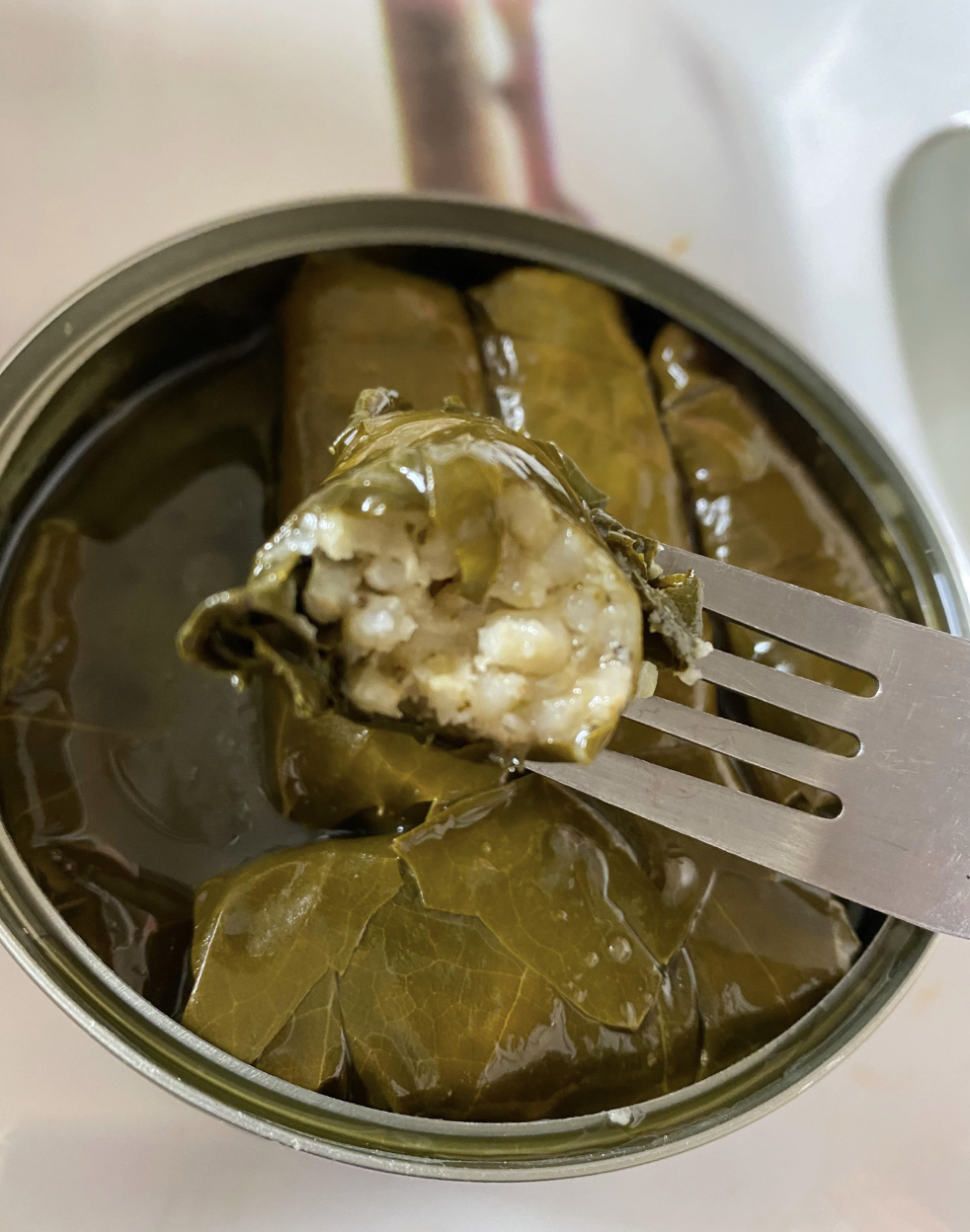 A fork holding a stuffed grape leaf with visible rice inside, over an open can filled with more stuffed grape leaves