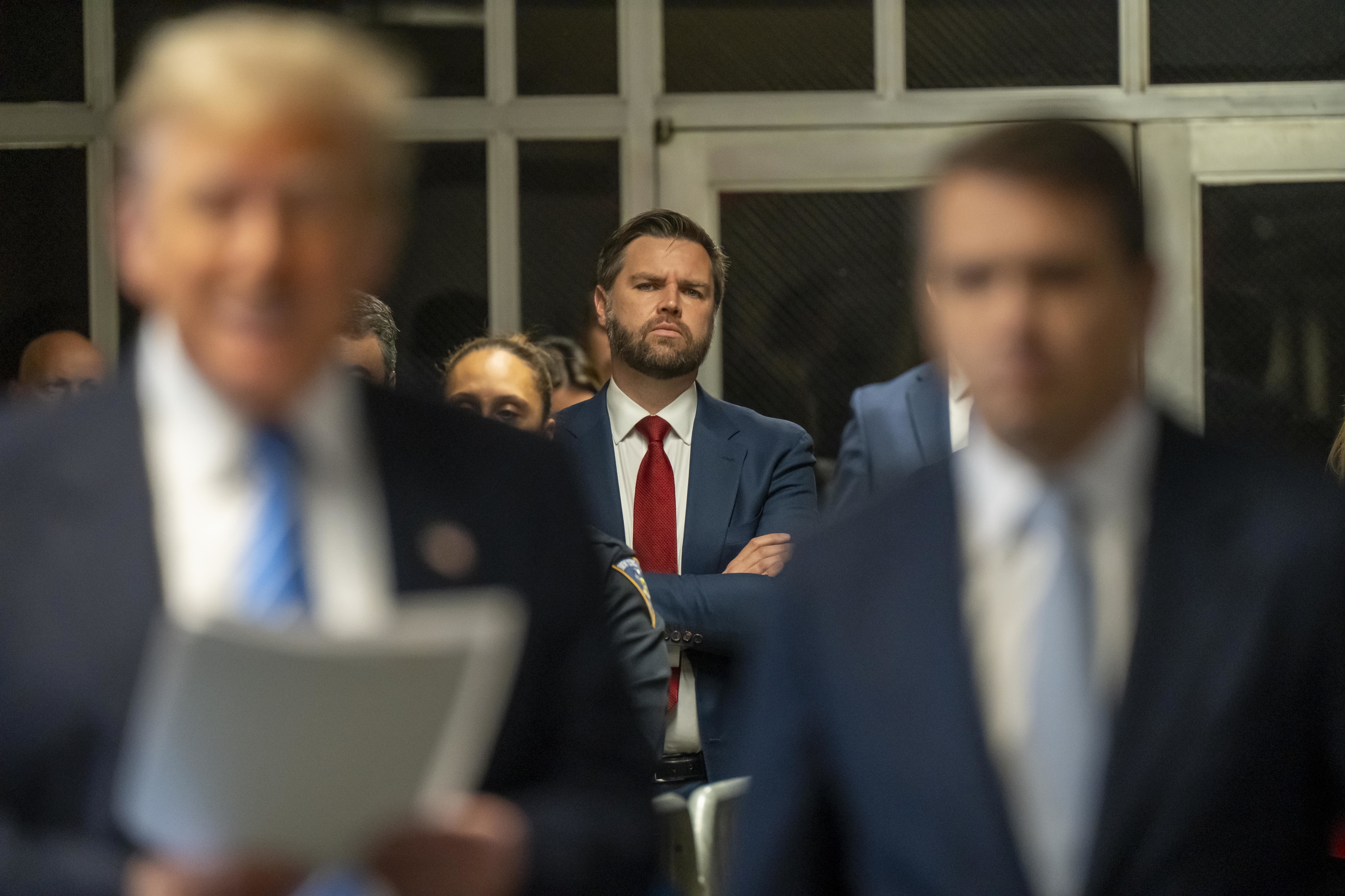 Donald Trump holds a speech in the foreground. A man in a blue suit and red tie stands with arms crossed in the background, looking forward
