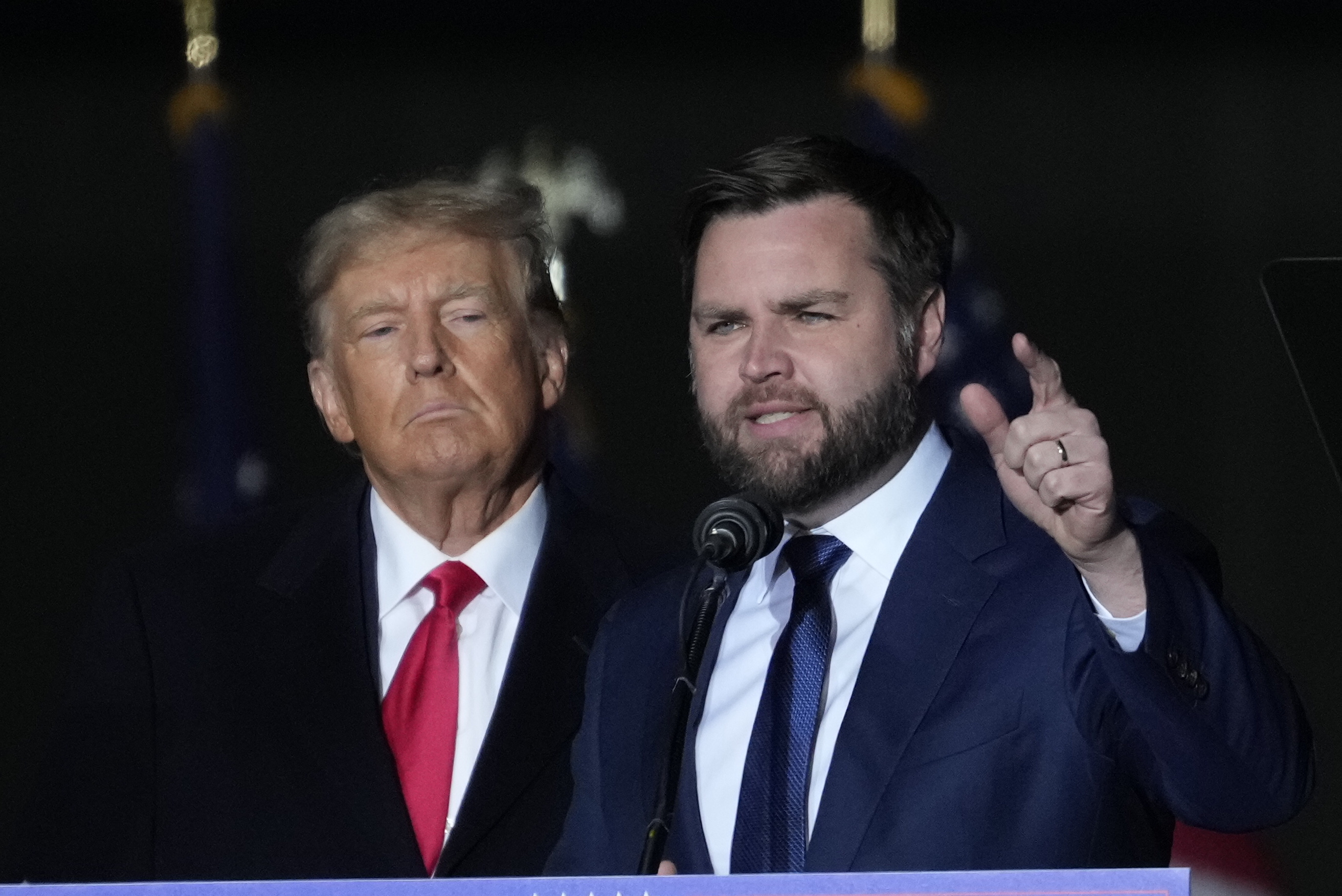 Donald Trump and J.D. Vance stand on stage. Trump is in a black suit with a red tie. Vance, gesturing while speaking, is in a blue suit with a white shirt and tie