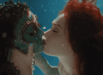 Two underwater characters kiss, one wearing aquatic-themed makeup and the other with flowing hair