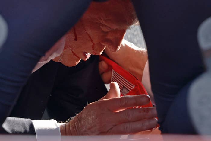 Former President Donald Trump is seen with blood on his face, wiping sweat from his chin as he kneels behind multiple individuals, with a focus on red cloth