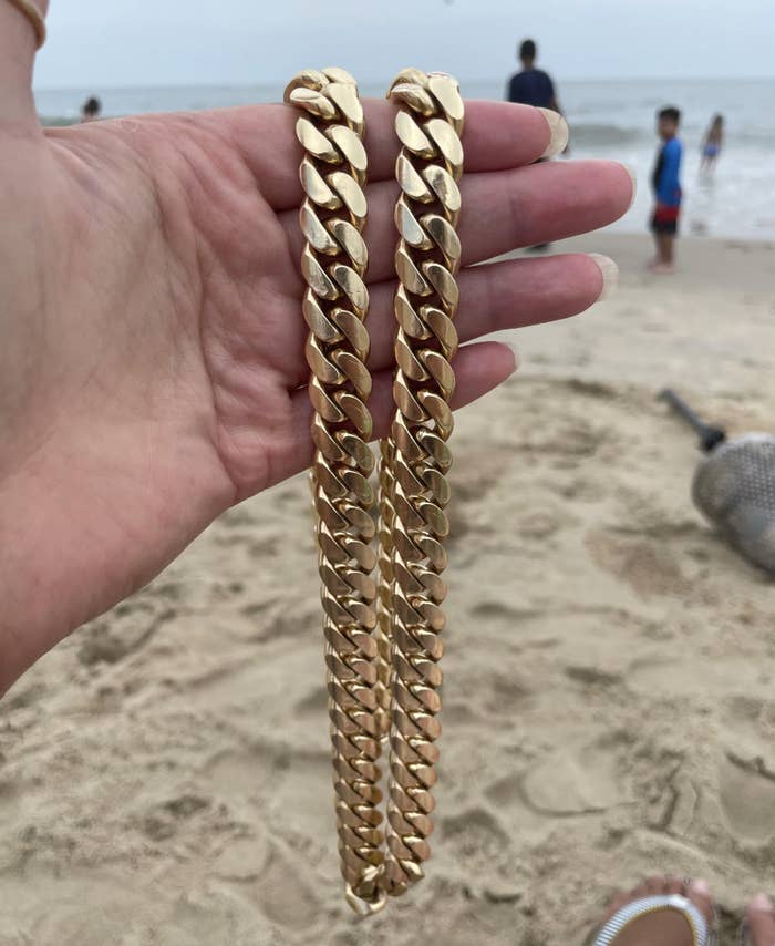 A person holds two gold chain necklaces in their hand at a beach. People and the ocean can be seen in the background