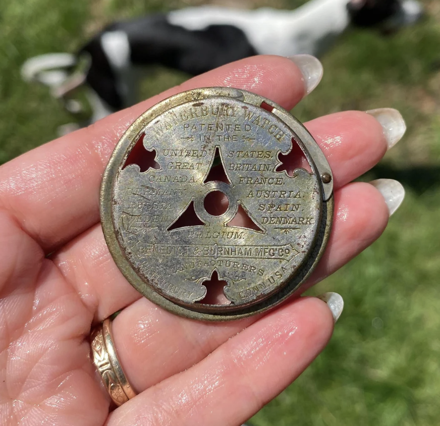 A hand holds a circular metal object labeled with various country names and "Burnham MFG CO," with a distant background showing part of a dog