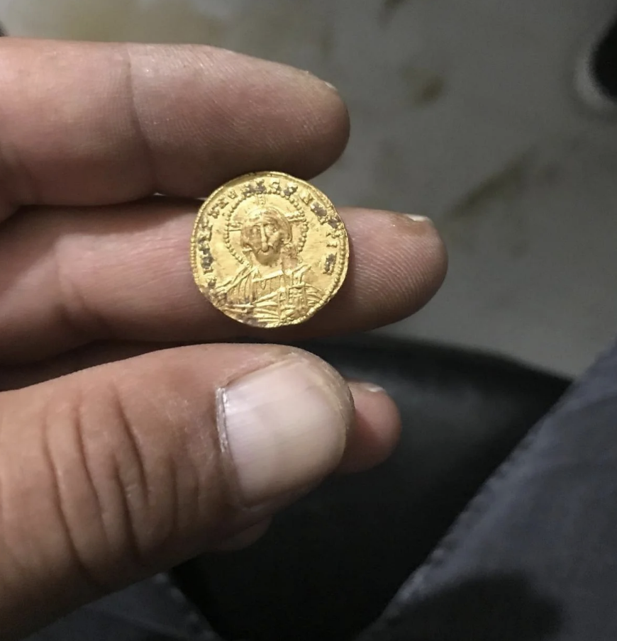 A hand holds a gold coin featuring an ancient figure and text. The coin has intricate designs and inscriptions. No individuals are present in the image