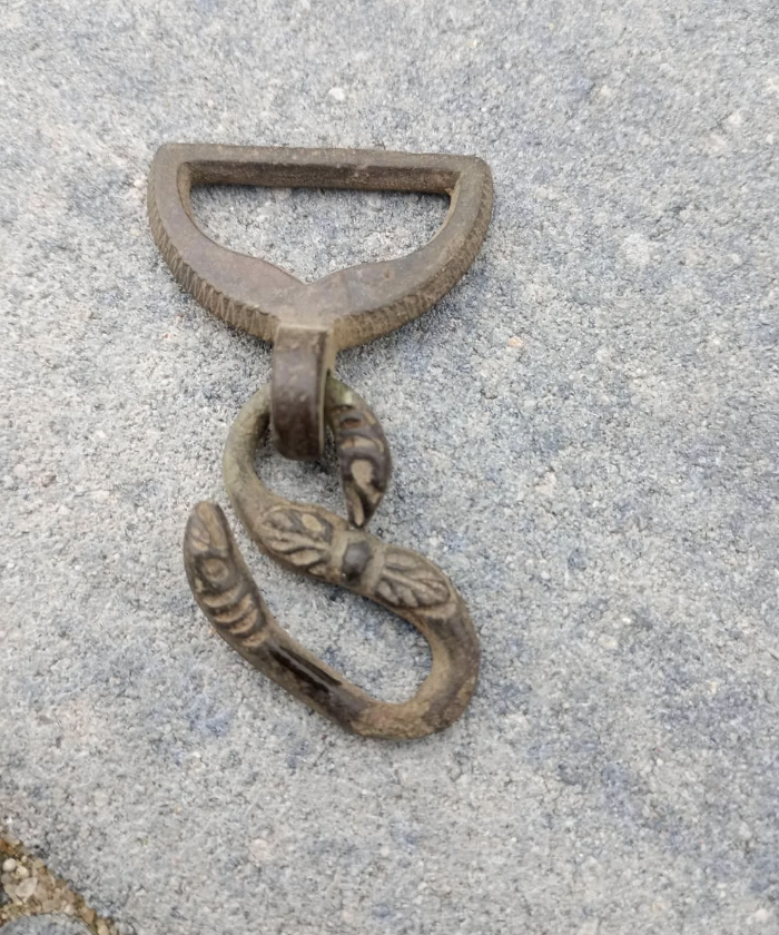A vintage, ornate metal belt buckle with an S-shaped design lying on a concrete surface