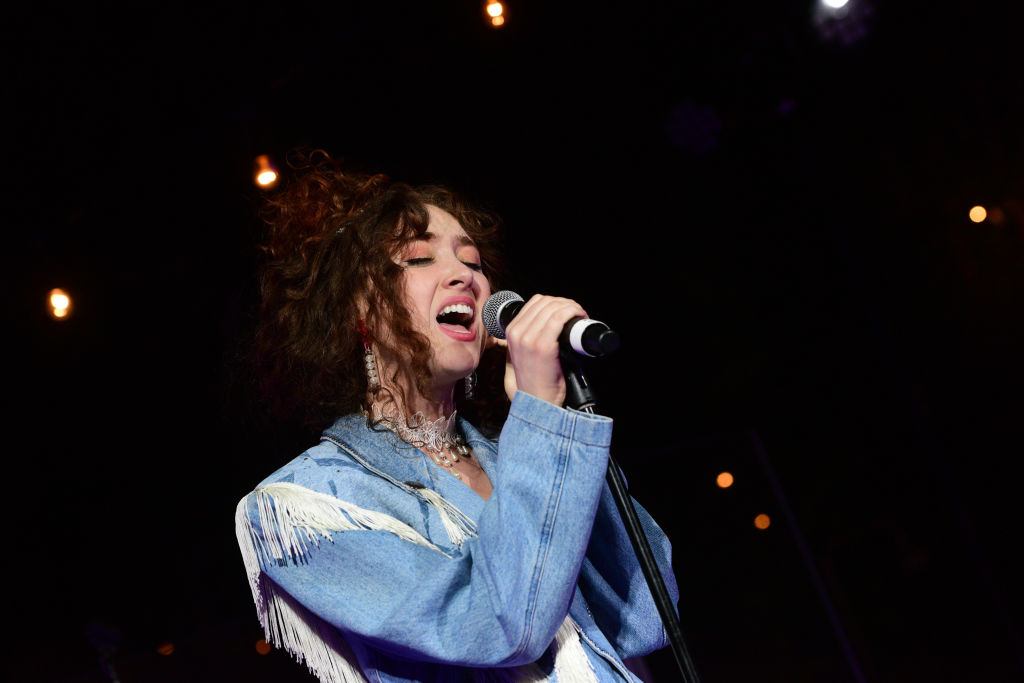 A person in a fringed jacket sings passionately into a microphone on stage