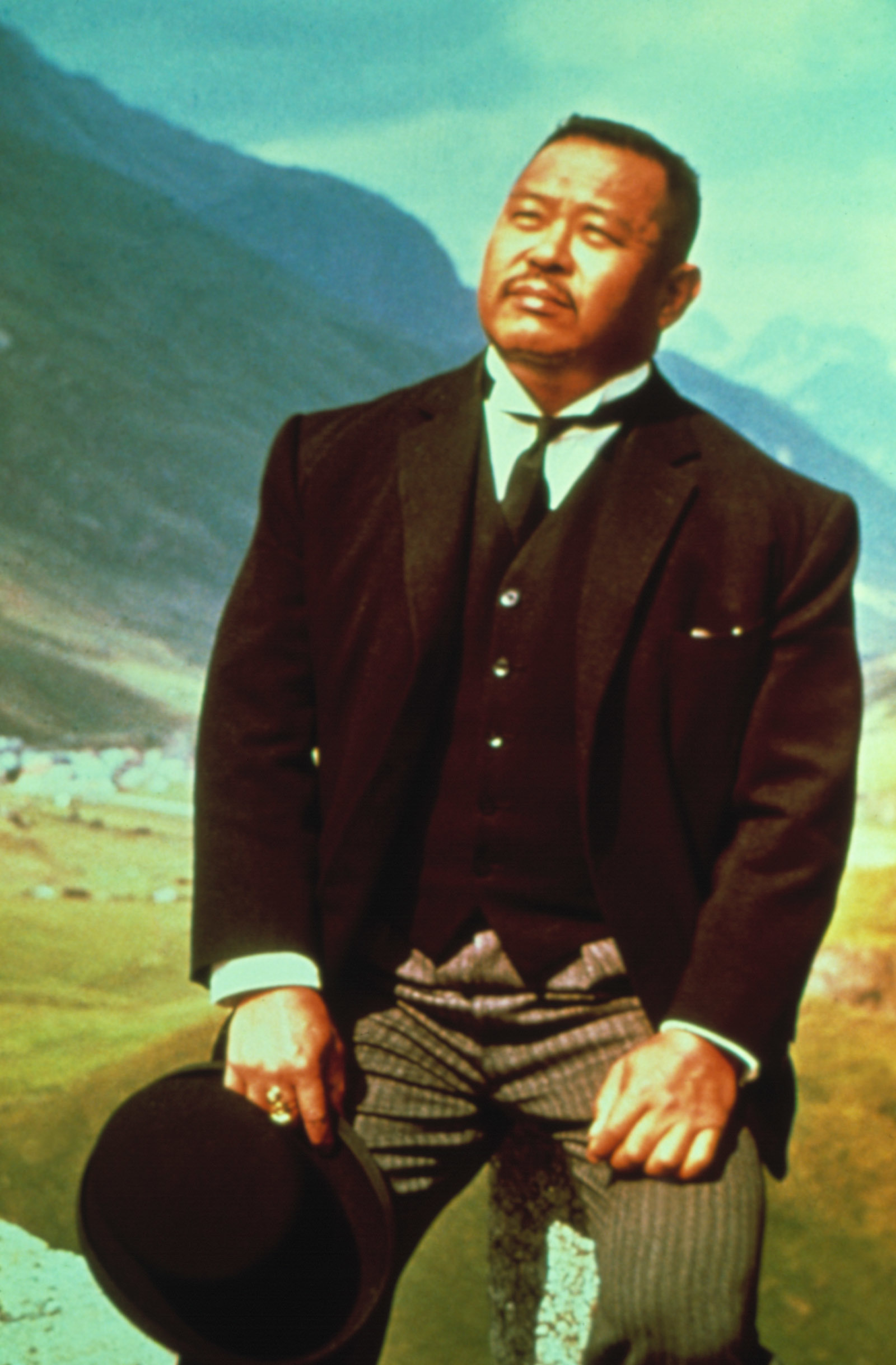 Oddjob stands outdoors in a mountainous region, wearing a formal suit and holding a hat