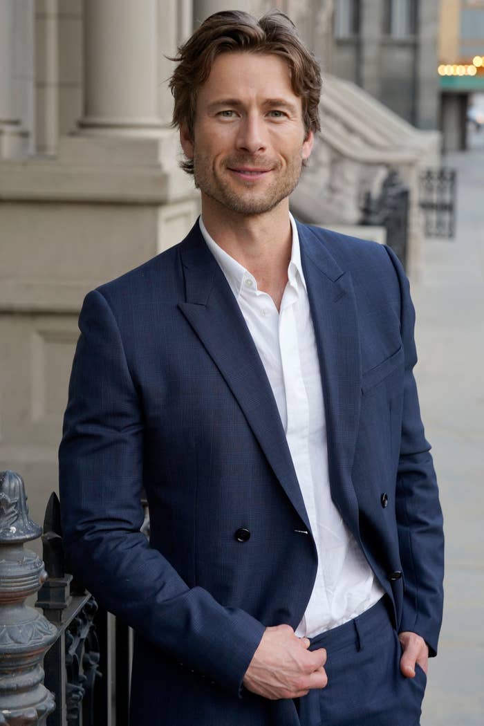 Glen Powell in a tailored suit stands outdoors, smiling with one hand in his pocket