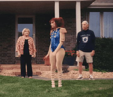 Woman in a blue sequin leotard with puffy shoulders and tall shoes dances on a lawn, while two older adults look on in the background