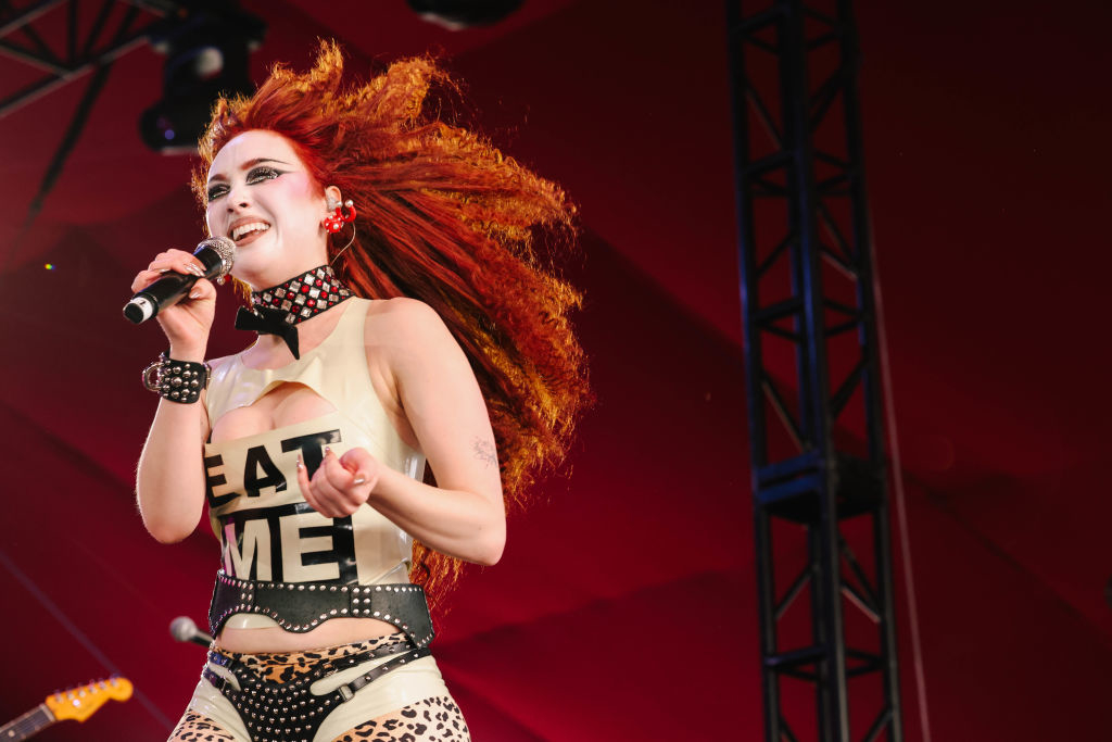 A singer with vibrant orange hair performs on stage, wearing a unique outfit with "EAT ME" written on it