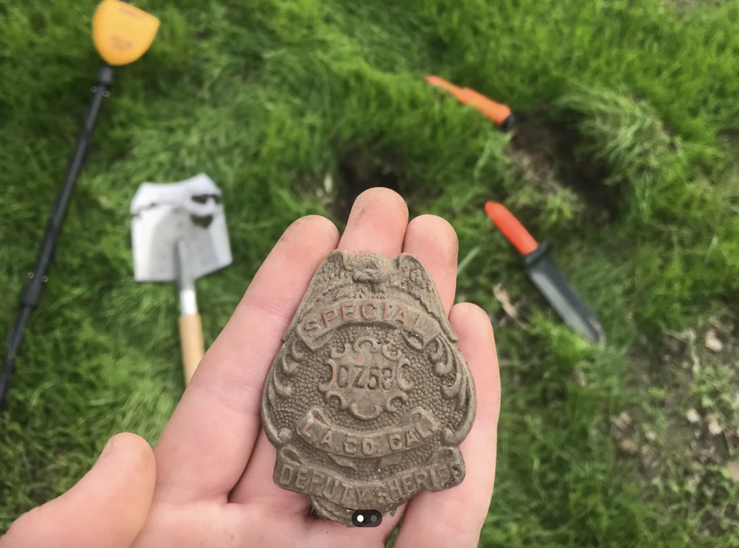 A hand holding an old "Special Deputy Sheriff" badge, with a shovel and metal detector on the grass in the background