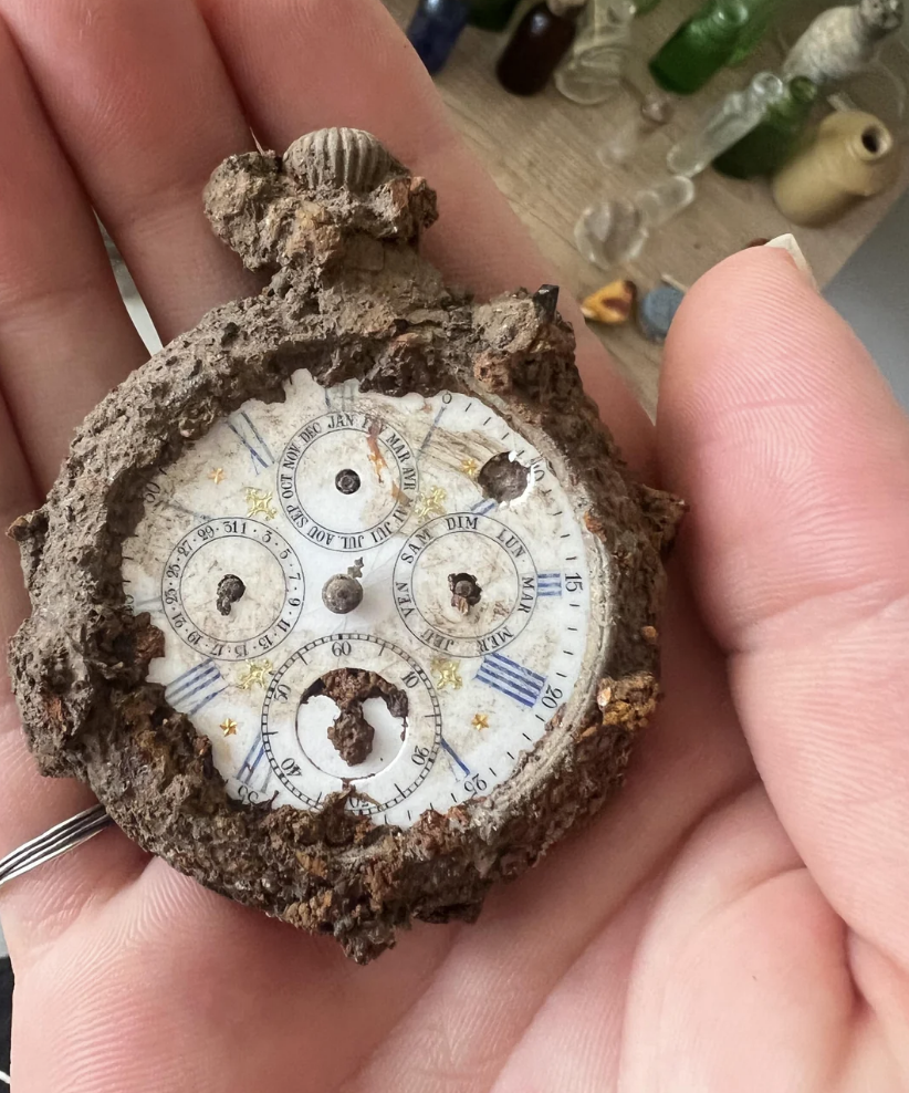 A hand holding a corroded antique pocket watch with multiple dials, showing Roman numerals, heavily encrusted with dirt. Background includes various small bottles on a shelf